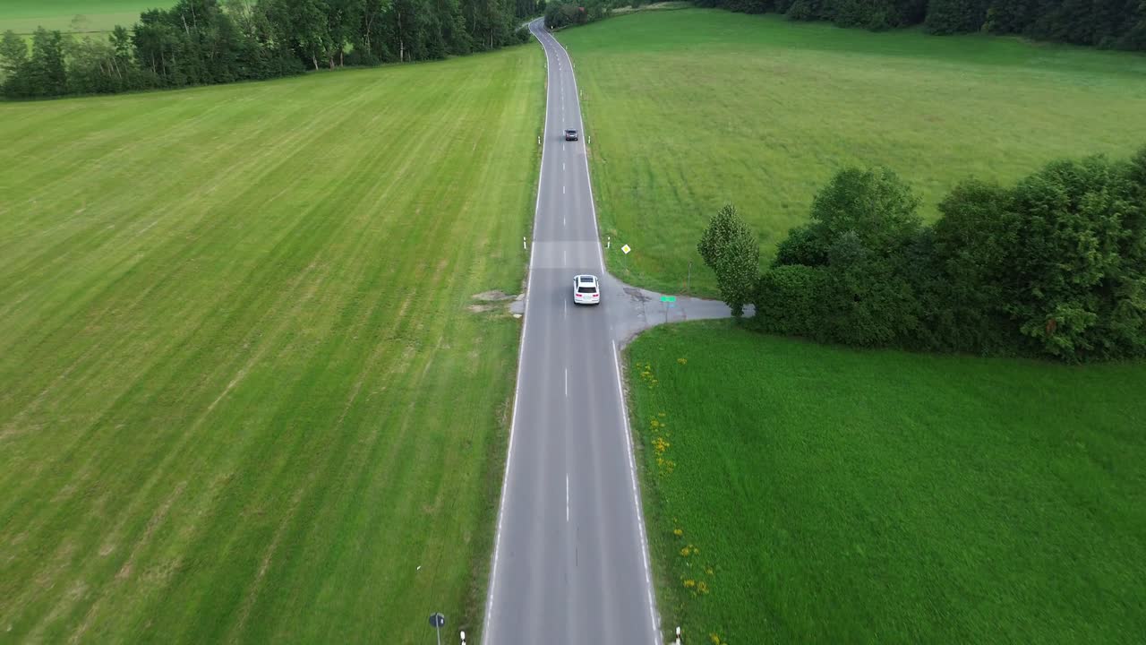Aerial drone view of two lane country road running through green fields with passing cars, forest edges, and peaceful rural landscape in foothills of Black Forest during daytime