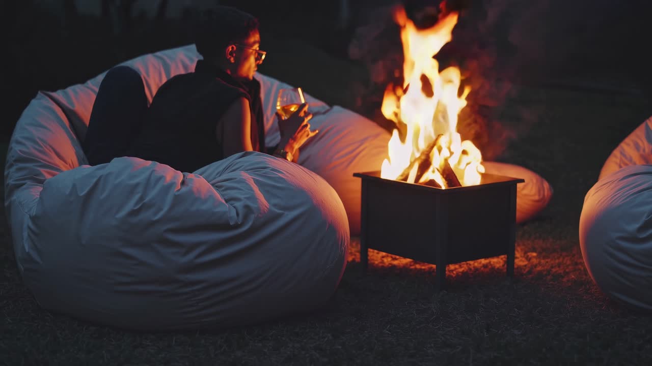 Man relaxing on a beanbag chair, sipping wine and enjoying the warmth of a cozy bonfire, surrounded by a tranquil outdoor ambiance filled with soft flames