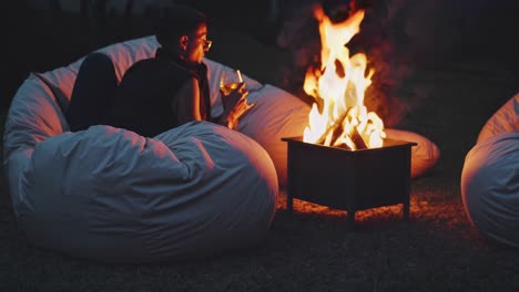 man relaxing on a beanbag chair, sipping wine and enjoying the warmth of a cozy bonfire, surrounded by a tranquil outdoor ambiance filled with soft flames