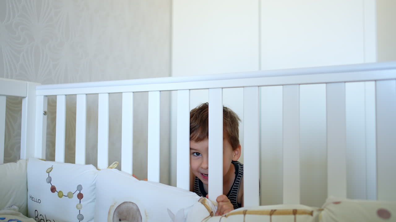 Lovely baby boy stands at his crib looking through the poles. Cute child plays hide-and-seek in the bedroom.
