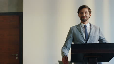 vista trasera de un hombre de negocios caucásico con ropa formal caminando entre la audiencia hacia el podio en una sala de conferencias, luego se gira y sonríe