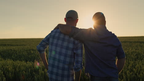 the farmer hugs his adult son and looks forward together to the wheat field where the sun is setting. family business and traditions concept