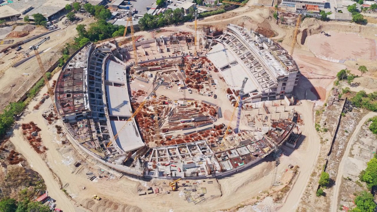 Grande estádio circular com estruturas de concreto e guindastes de torre durante a fase de construção, mostrando o progresso contínuo do enorme projeto arquitetônico de uma perspectiva aérea de cima para baixo