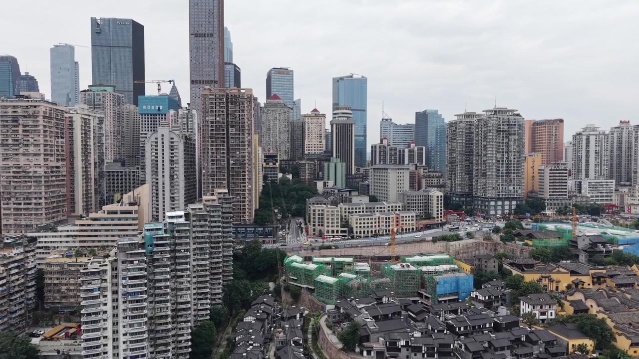 Panorâmica aérea lenta sobre um bairro tradicional com telhados escuros, cercado por arranha-céus modernos, revelando o denso horizonte do CBD de Chongqing ao fundo, céu nublado, Chongqing, China.