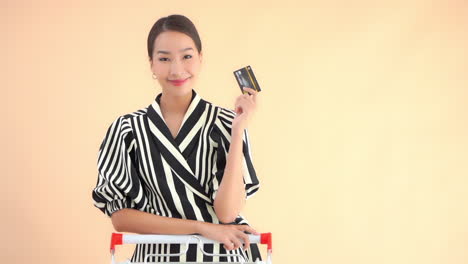 A-pretty-young-woman-leans-on-the-handle-of-a-grocery-cart-raises-her-hand-holding-a-credit-card