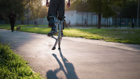 un tipo caminando con piernas protésicas cargadas de muelles al aire libre en un parque soleado con vegetación y camino pavimentado, con pantalones oscuros. las sombras se proyectan en el camino, capturando el movimiento