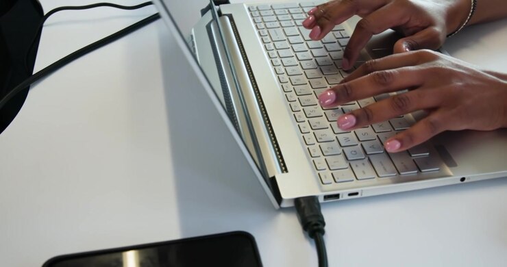 Weibliche Hände tippen, nachdem die Finger Tasten auf einem silbernen Laptop an einem weißen Schreibtisch für die Arbeit gedrückt haben.