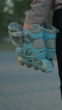 una chica, vestida con una chaqueta de melocotón y pantalones negros, lleva un par de patines y una bolsa de gimnasia mientras camina por un parque durante la puesta de sol. centrándose en los patines y su vestimenta casual