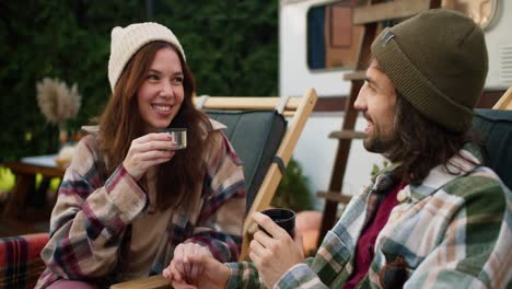 Close-up-shot-of-a-happy-brunette-girl-drinking-tea-from-a-thermos-mug-together-with-her-brunette-boyfriend-in-a-green-checkered-shirt-who-holds-her-hand-and-communicates-with-her-during-his-picnic-outside-the-city-near-a-trailer-on-chairs