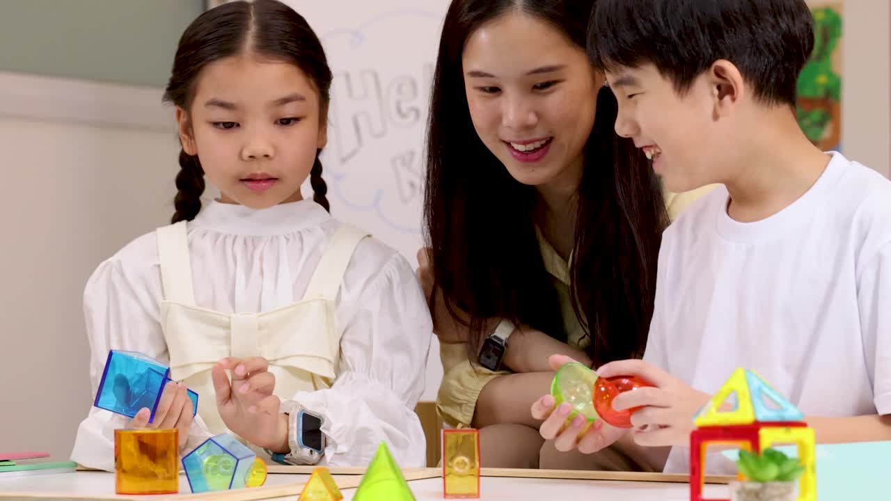 明るい教室で、2人の子供と先生がブロックを使ってカラフルな幾何学的な形を作っています。
