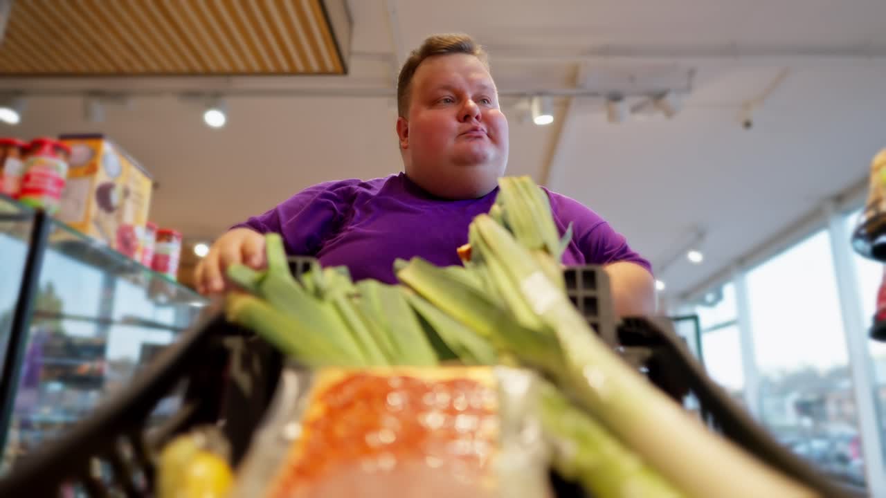 View from the cart side: A happy overweight man wearing a purple T-shirt walks through the supermarket, looks at the products. Shallots are already in the man's cart