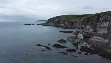 Aerial-Drone-Shot-of-Ayrmer-Cove-in-Devon,-England-with-calm-crystal-blue-waters-and-stunning-cliffs-,-rolling-green-countryside-