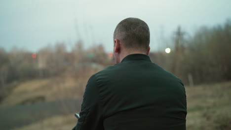 a close view of a man in a green jacket sitting down, captured from behind, looking out over a blurred, forested landscape at dusk, with a distant light visible in the background