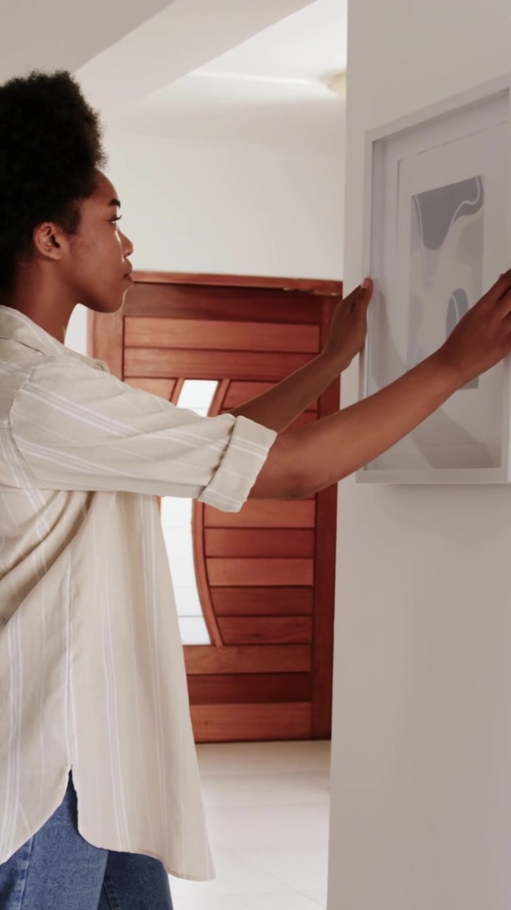 Video verticale: Donna con top beige che porta delle stampe, allinea e appende una cornice vicino alla porta, con spazio per il testo.
