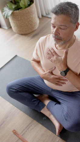 vertical video of senior biracial man sitting practicing yoga at home, slow motion