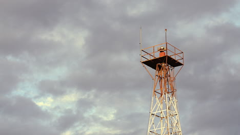 a rotating beacon light, mounted on a tower at a small airport, is shown in the evening, set against a strong clouds