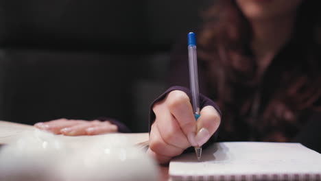 close up view of female hand with acrylic nails writing in spiral notebook using pen while holding open book on wooden table. white decorative elements on table and blurred background