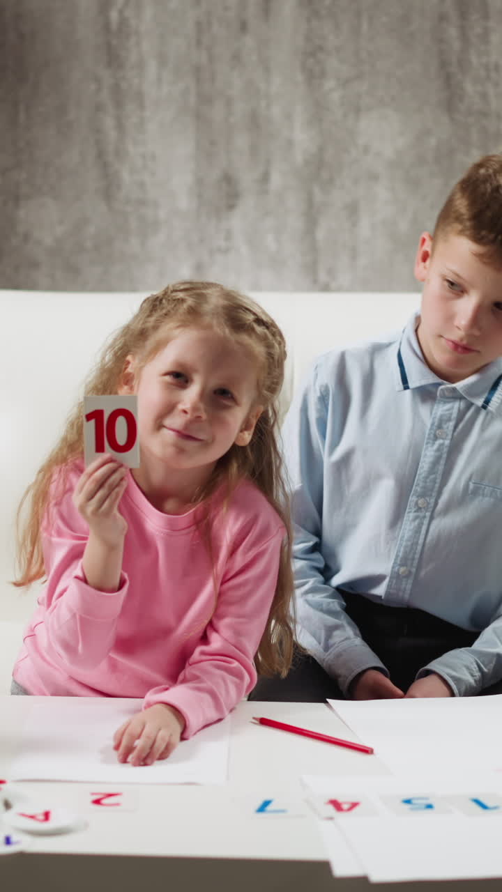 Smiling long haired schoolgirl in pink sweatshirt shows card with digit ten to camera and boy looks at classmate on sofa at remote lesson at home slow motion