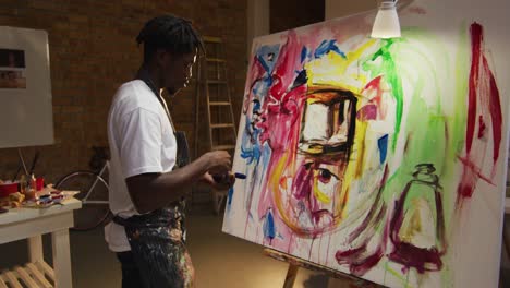 African-american-male-artist-wearing-apron-painting-with-palette-knife-on-canvas-at-art-studio