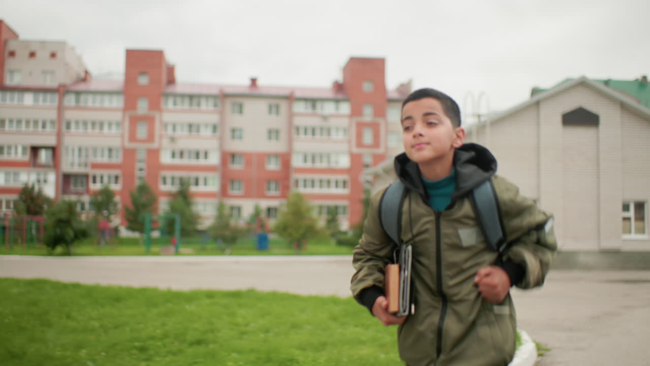 밝은 미소를 지으며 팔에 메모장을 끼고 배낭을 멘 아이가 초록빛 식물과 붉은 벽돌의 주거 구역이 배경으로 보이는 대학 건물 근처에서 뛰고 있어요. 명랑한 학업 분위기가 느껴지네요.