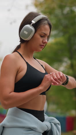 side view of a woman wearing black sportswear with blue hoodie tied around her waist, walking and checking time on wristwatch while listening to music with headphones in a park