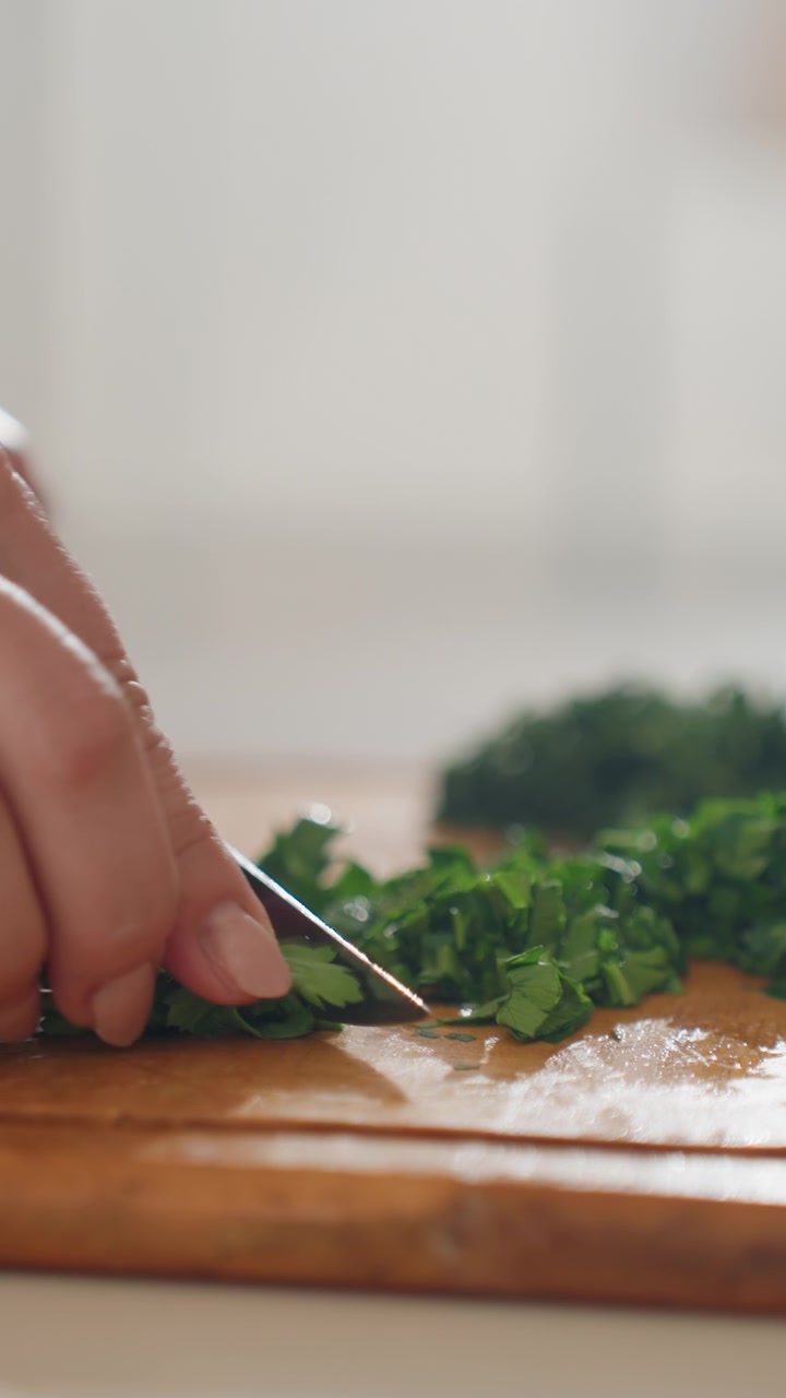 La mano sinistra trita erbe fresche verdi su un caldo tagliere di legno, le punte delle dita arricciate per tenere il mazzetto mentre un piccolo coltello affetta e trita le foglie; visibile un movimento di taglio verso il basso con erbe sparse.