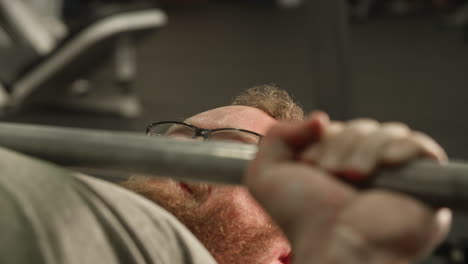 Sweating-man-with-glasses-bench-pressing-inside-of-gym,-Close-Up