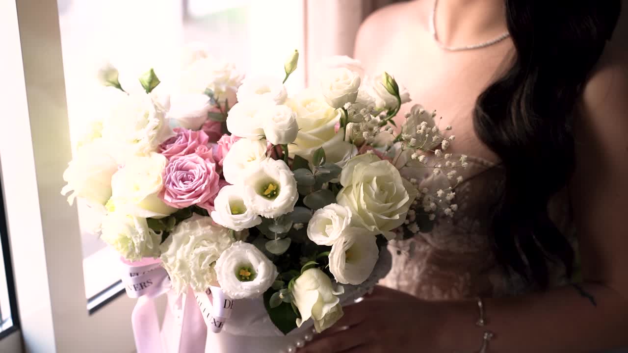 Una donna sta in piedi vicino a una grande finestra tenendo un bouquet di fiori. Si prepara per un evento speciale. Il bouquet contiene diversi tipi di fiori.