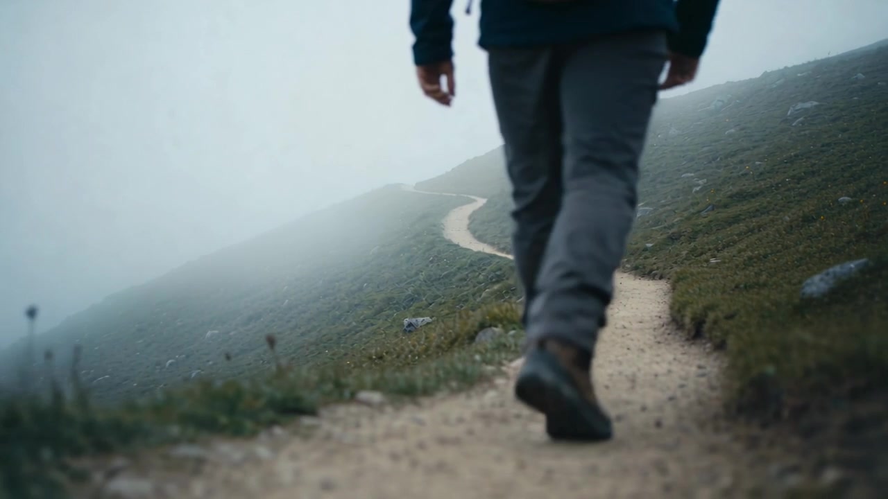Telecamera posteriore bassa che segue un singolo camminatore che avanza lungo un sentiero nebbioso, dirigendosi nella nebbia, indossando stivali.