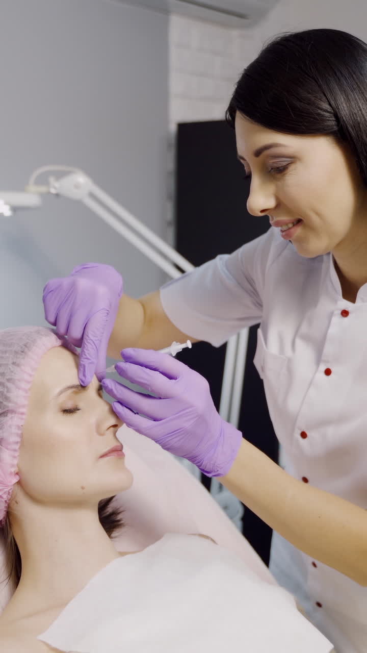 A pretty cosmetologist with a dark hair color makes Botox for a woman who lays on the couch in a beauty salon. Botulinum toxin Vertical video