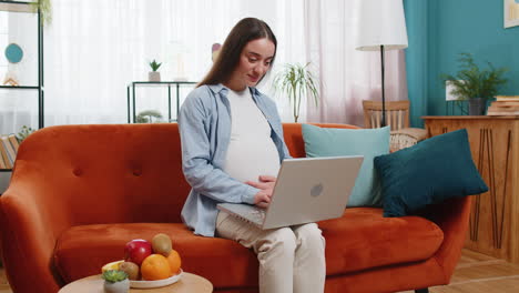 happy pregnant woman touching belly while using working on laptop computer sitting on home sofa