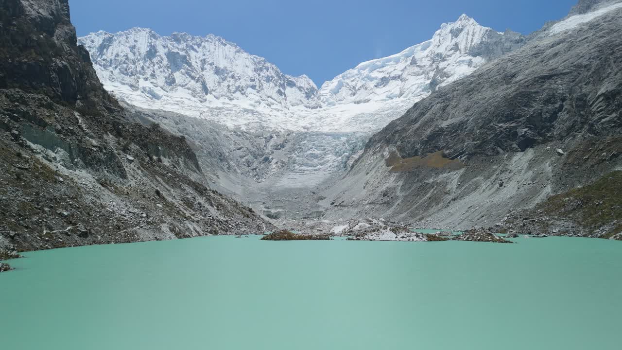 A stunning aerial trucking shot from the right, revealing turquoise Laguna Llaca, rocky moraines, and snow-capped peaks in Peru’s Cordillera Blanca