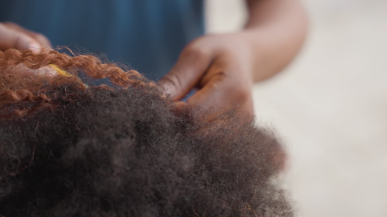 Primo piano di mani che intrecciano capelli naturali, le dita dello stilista intrecciano una treccia stretta lungo riccioli e ciocche testurizzate, scena intima del salone con luce naturale soffusa, movimento lento e focalizzato, particelle di polvere visibili.