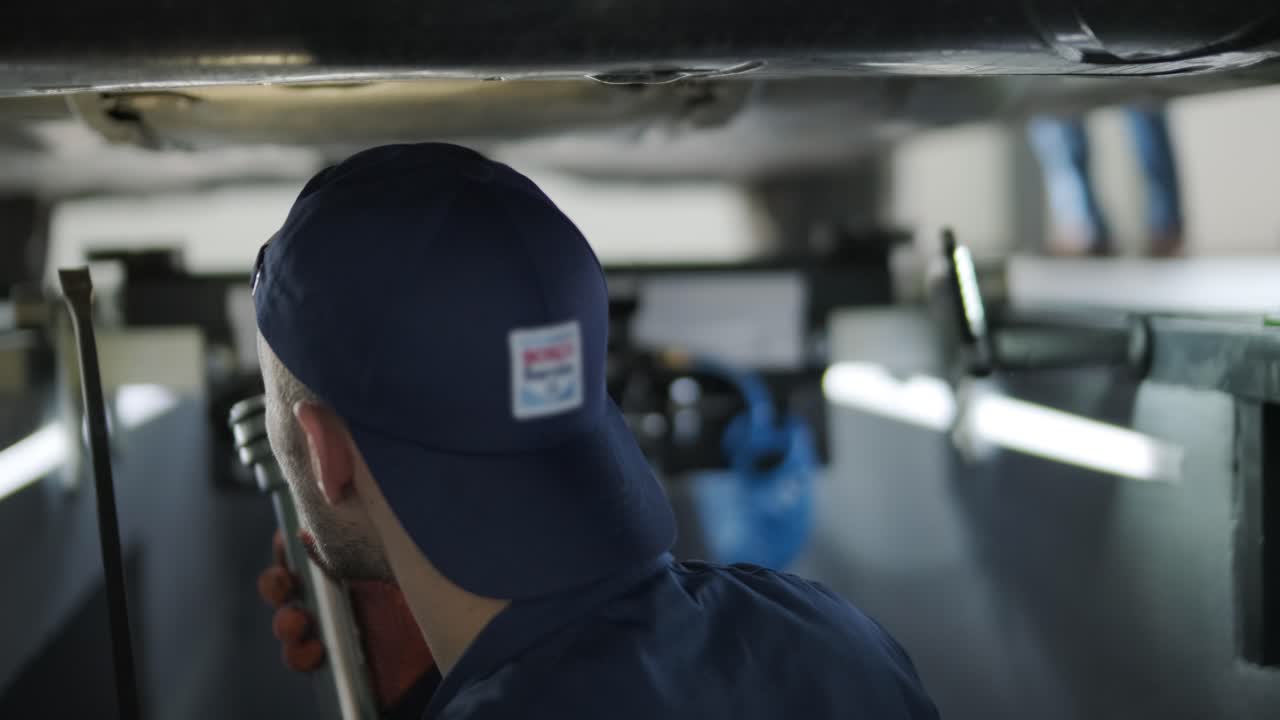 mecánico en uniforme reparando la parte inferior de un coche usando herramientas.