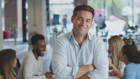 portrait of businessman with colleagues in background sitting around table in open plan office