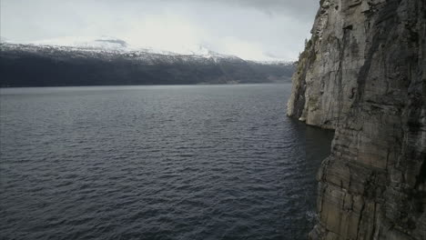 flying over norwegian fiord close to rocky shore, mountains with snow in background, aerial shot