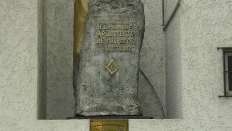 a stone monument with the masonic symbols of the square, the compass and the letter g and above a phrase in spanish translated to "man, architect of his own destiny"