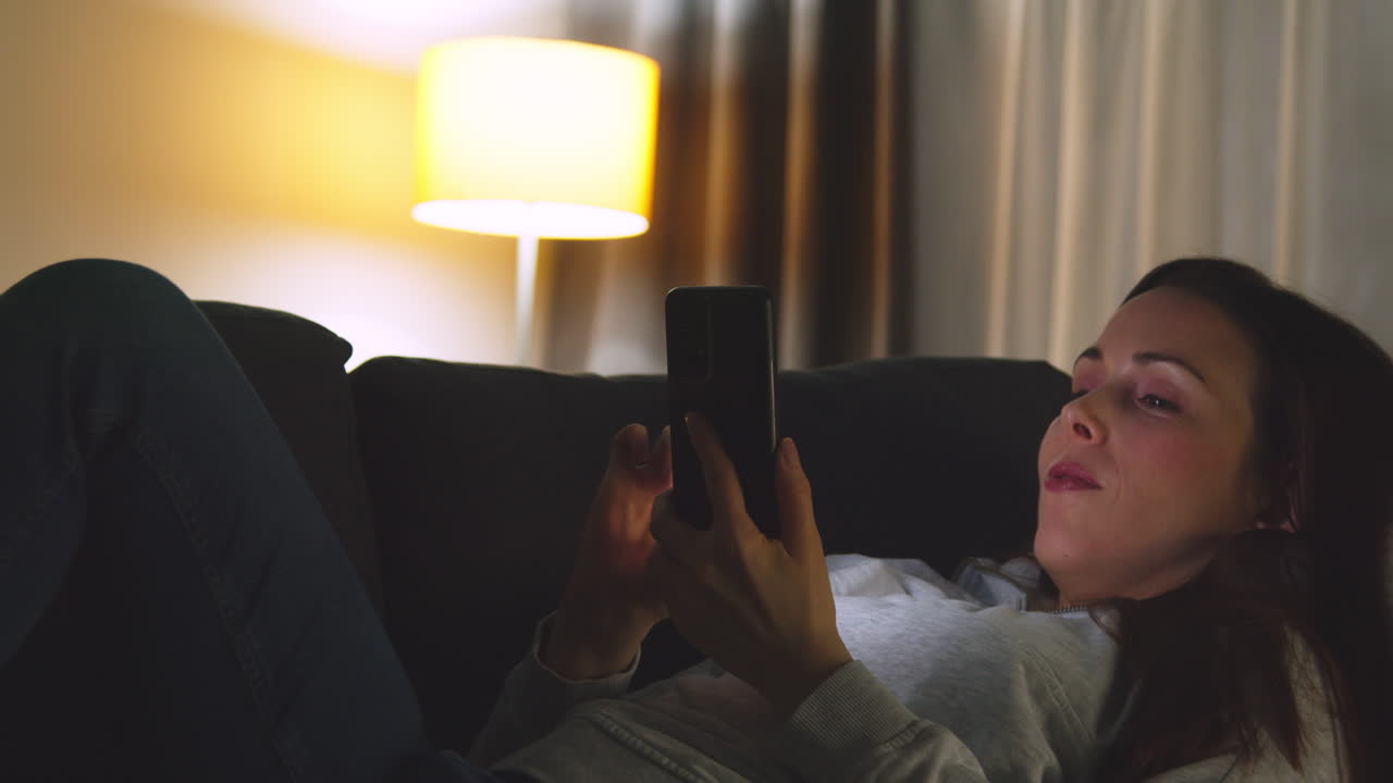 밤에 집에서 소파에 누워서 휴대 전화로 온라인 콘텐츠를 스트리밍하거나 보는 여성