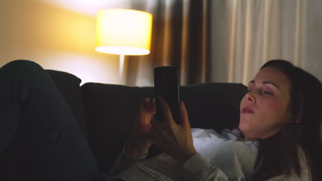 woman lying on sofa at home at night streaming or looking at online content on mobile phone 9