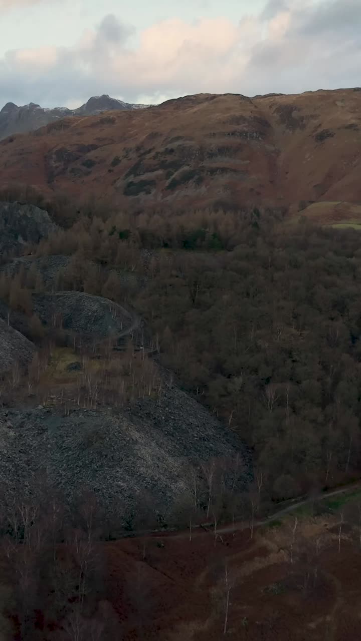 Vertical forward drone flight over slate mounds and winter trees toward hilltop peaks and fields. Heavy clouds darken the blue sky, adding a moody, foreboding atmosphere. Strong establishing shot