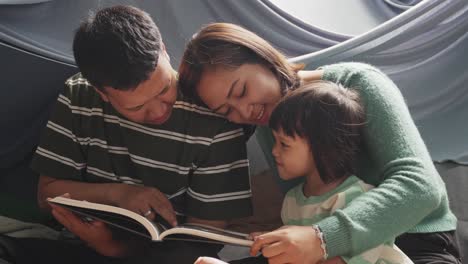 Padre-Y-Madre-Asiáticos-Felices-Leyendo-Un-Libro-A-Su-Pequeña-Hija-En-Casa