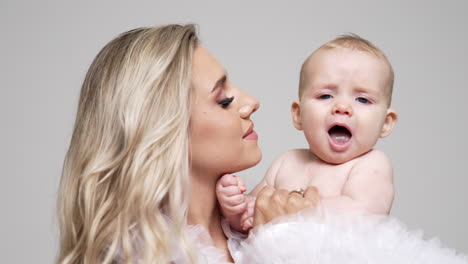 beautiful blonde woman kissing her lovely baby boy on the cheek. mother with her infant son in studio. white backdrop.