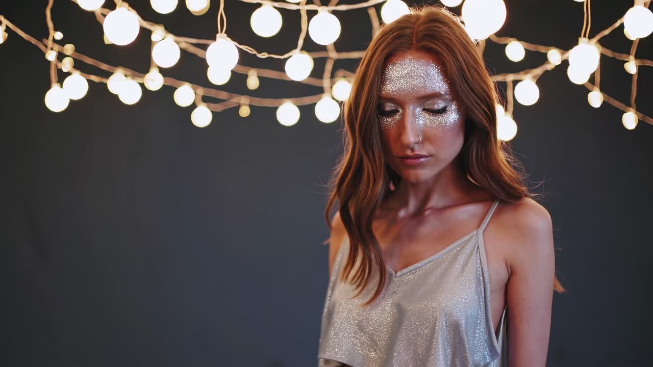 Blurred fashion model posing gracefully under glowing string lights, showcasing striking silver makeup and a stylish two piece outfit in a serene studio with a dark backdrop