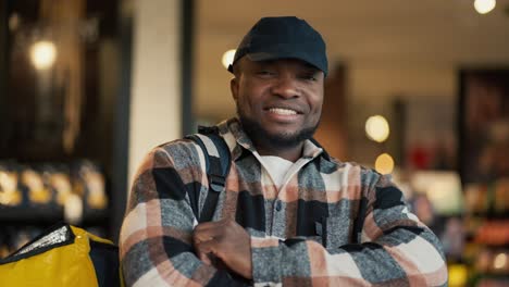 Retrato-De-Un-Hombre-Feliz-De-Piel-Negra-Con-Una-Camisa-A-Cuadros-Y-Una-Gorra-Negra-Que-Trabaja-Como-Repartidor-Y-Lleva-Una-Gran-Bolsa-Amarilla-Sobre-Sus-Hombros-Mientras-Busca-Los-Productos-Necesarios-En-Un-Supermercado-Moderno.