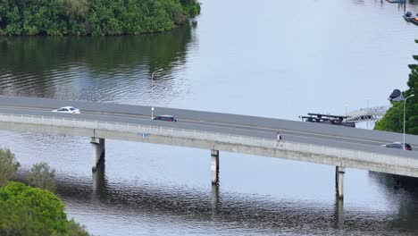vehicles and pedestrians traverse a bridge over calm water, surrounded by greenery, under clear daylight