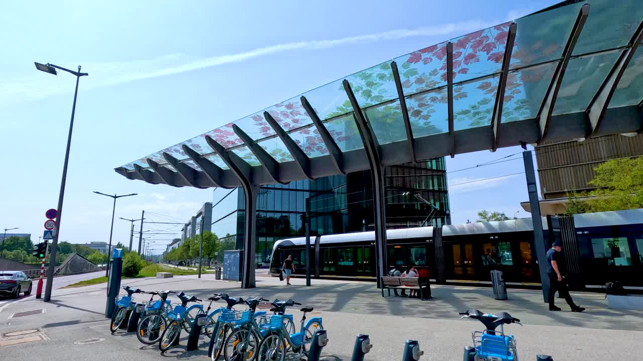 A modern tram approaches and stops at a contemporary glass-covered train station in Luxembourg, with bike rentals and urban buildings under bright daylight