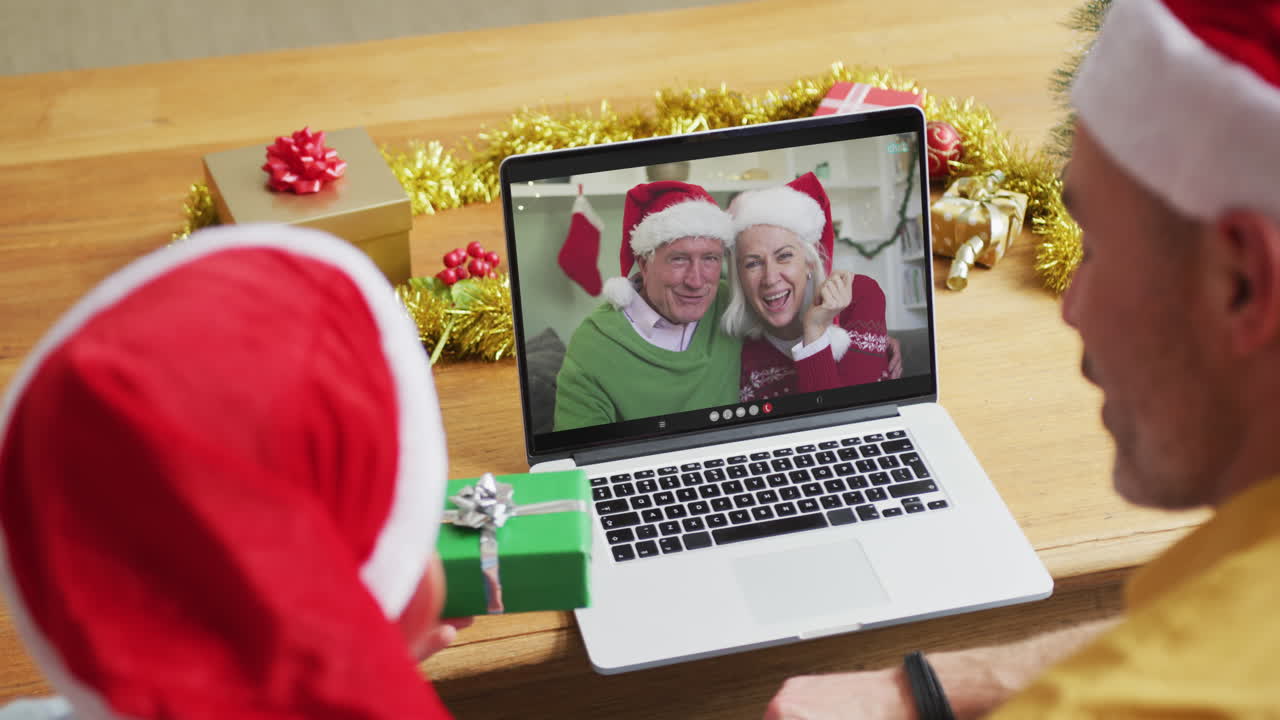 padre y hijo caucásicos con sombreros de santa usando una computadora portátil para una videollamada de navidad con una pareja en la pantalla