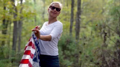 pretty blonde woman with sunglasses on in a forest spinning around to the right with a us flag flying