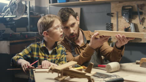 cheerful young caucasian carpenter working at desk in workshop while his small son is playing next to him