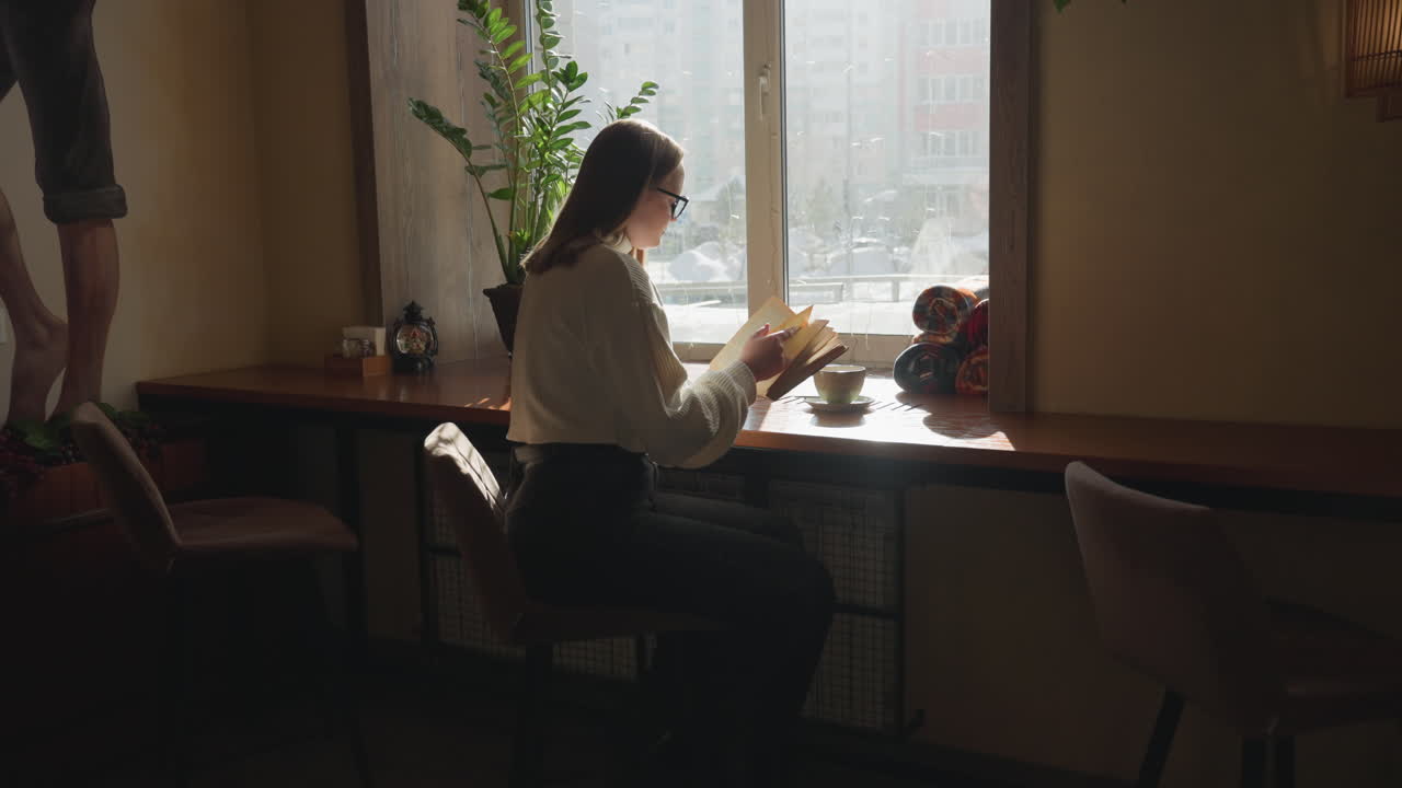 High school girl sits near bright window reading novel with focus, flipping page as sunlight pours over wooden table with mug and rolled blanket, indoor plants and mural adding warm atmosphere